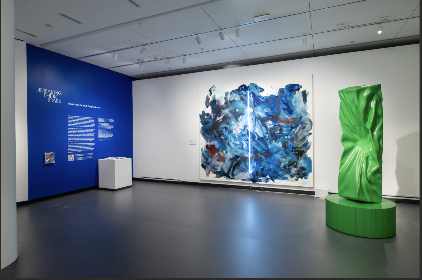 A gallery view of a space with white and blue walls. An abstract blue painting is on the center wall, with a large green metal sculpture to the right.